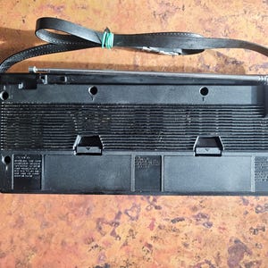 Puede incluir: Radio vintage negra con correa de cuero y antena telesc&oacute;pica. La radio tiene forma rectangular con rejilla de altavoz y varios paneles de control. Una goma el&aacute;stica verde asegura la correa.