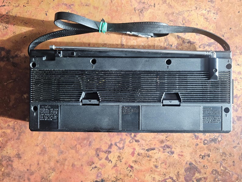 Puede incluir: Radio vintage negra con correa de cuero y antena telesc&oacute;pica. La radio tiene forma rectangular con rejilla de altavoz y varios paneles de control. Una goma el&aacute;stica verde asegura la correa.