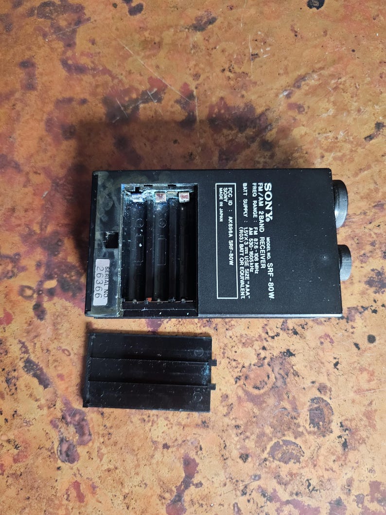 Puede incluir: Receptor de 2 bandas FM/AM Sony SRF-80W negro con compartimento de bater&iacute;a y tapa desmontada. La radio tiene dos perillas de sintonizaci&oacute;n y un n&uacute;mero de serie 26366. La radio est&aacute; fabricada en Jap&oacute;n.