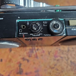 Puede incluir: Primer plano del panel de control de un boombox negro vintage. Incluye perillas de volumen y sintonizaci&oacute;n, un bot&oacute;n de expulsi&oacute;n y una entrada est&eacute;reo FM. El boombox tiene un asa y rejillas de altavoz.