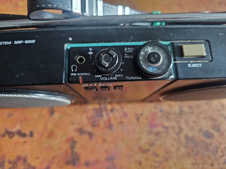 Puede incluir: Primer plano del panel de control de un boombox negro vintage. Incluye perillas de volumen y sintonizaci&oacute;n, un bot&oacute;n de expulsi&oacute;n y una entrada est&eacute;reo FM. El boombox tiene un asa y rejillas de altavoz.