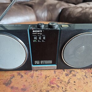 Puede incluir: Radio est&eacute;reo FM Sony negra con rejillas de altavoz plateadas y una antena retr&aacute;ctil. La radio tiene un panel de control negro con el texto "FM STEREO" y el logotipo de Sony. La radio est&aacute; sobre una superficie marr&oacute;n.