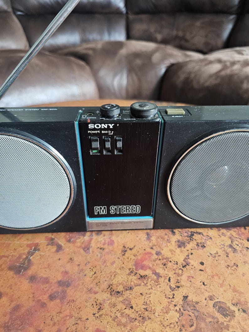 Puede incluir: Radio est&eacute;reo FM Sony negra con rejillas de altavoz plateadas y una antena retr&aacute;ctil. La radio tiene un panel de control negro con el texto "FM STEREO" y el logotipo de Sony. La radio est&aacute; sobre una superficie marr&oacute;n.