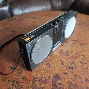Puede incluir: Una radio port&aacute;til negra Sony ICF-5900W con una rejilla de altavoz plateada y un altavoz blanco. La radio tiene una correa negra adjunta y varios botones. El texto "SONY" es visible en la radio.