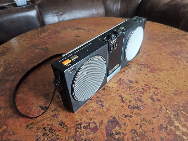 Puede incluir: Una radio port&aacute;til negra Sony ICF-5900W con una rejilla de altavoz plateada y un altavoz blanco. La radio tiene una correa negra adjunta y varios botones. El texto "SONY" es visible en la radio.