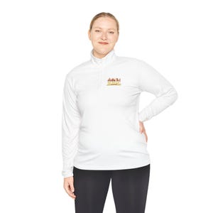 Könnte beinhalten: Ein weißer Langarm-Pullover mit Viertelreißverschluss und einer kleinen, farbenfrohen Grafik auf der linken Brust. Die Grafik zeigt eine stilisierte Häuserreihe in Orange-, Rot- und Gelbtönen mit dem Schriftzug "DURHAM" in goldener Schrift.