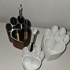 Puede incluir: Tres organizadores de escritorio con forma de pata. Uno es blanco, otro tiene un degradado de colores y el tercero es blanco. Cada uno tiene un centro en forma de corazón. Un bolígrafo está colocado en dos de los organizadores.