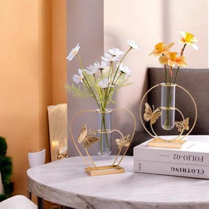 Può includere: Due vasi decorativi in metallo dorato con tubi di vetro trasparente per fiori. Un vaso è a forma di cuore con decorazioni a farfalla, l'altro è un cerchio con decorazioni a farfalla. Fiori bianchi e gialli nei vasi.
