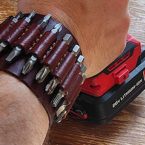 Könnte beinhalten: Ein braunes Lederarmband mit mehreren Schraubendreherbits. Das Armband wird am Handgelenk einer Person neben einem roten und schwarzen 20V Lithium-Ionen-Akku getragen.