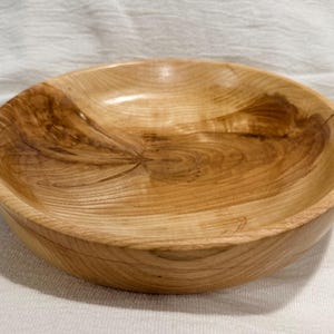 May include: A shallow, round wooden bowl featuring a natural light brown finish with prominent dark wood grain patterns, swirling textures, and a smooth, polished surface that highlights the organic characteristics of the timber material.
