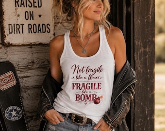 May include: A white racerback tank top featuring the maroon text Not fragile like a flower, FRAGILE like a BOMB, with a small graphic of a round bomb with a lit fuse. The tank top is worn with a black leather jacket and blue denim shorts.