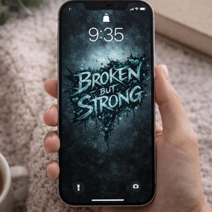 May include: A smartphone displays a wallpaper with the words "BROKEN BUT STRONG" in a teal, distressed font against a dark, textured background. The time 9:35 is visible at the top. The phone is held in a hand.