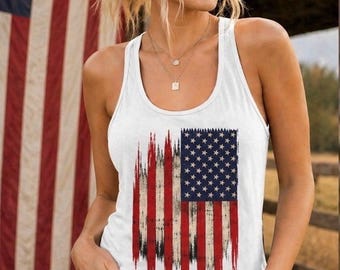 May include: White tank top featuring a distressed American flag design. The top has a scoop neck and is paired with denim shorts. The background includes an American flag and a wooden fence.