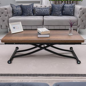 May include: A rectangular, brown wooden coffee table with a black metal base. The table is in front of a gray sofa with patterned pillows. The table has a closed size of 24x47 inches and an open size of 47x47 inches. The height is adjustable.