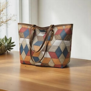 Könnte beinhalten: Eine farbenfrohe Tragetasche mit einem geometrischen Muster in Rot-, Blau-, Orange- und Beigetönen. Die Tasche hat braune Ledergriffe und -verzierungen und steht auf einer Holzoberfläche. Ein modisches Accessoire.