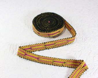 May include: A roll of decorative trim featuring a woven pattern in yellow, pink, and black. The trim is rolled up, with a portion unrolled to show the repeating design. The trim has a beaded edge.