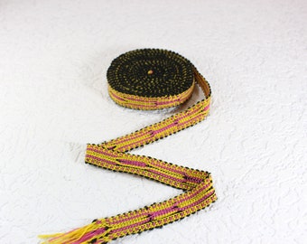 May include: A roll of decorative trim with a black, yellow, and pink woven pattern. The trim is unrolled, revealing a section with a fringed end. The trim is likely used for sewing, crafting, or embellishing various items.