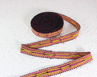 May include: A roll of decorative trim with a repeating pattern. The trim features a black base with pink and yellow woven details. The edge is finished with a black beaded trim. The trim is unrolled on a white surface.