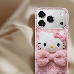 Może przedstawiać: R&oacute;żowe etui na telefon z tr&oacute;jwymiarowym wzorem Hello Kitty. Etui ma puszystą fakturę z białą twarzą Hello Kitty, r&oacute;żową kokardą i r&oacute;żowym wzorem kokardek. Etui ma przezroczystą obw&oacute;dkę.