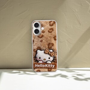 Może przedstawiać: Etui na telefon z motywem Hello Kitty. Etui ma brązowe i beżowe tło z nadrukiem w cętki i pluszową fakturę. Postać Hello Kitty znajduje się na środku, a pod nią napis "HelloKitty".