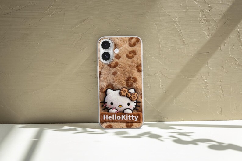 Może przedstawiać: Etui na telefon z motywem Hello Kitty. Etui ma brązowe i beżowe tło z nadrukiem w cętki i pluszową fakturę. Postać Hello Kitty znajduje się na środku, a pod nią napis "HelloKitty".