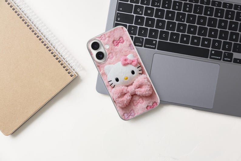 Może przedstawiać: R&oacute;żowe etui na telefon z białym kotem z kresk&oacute;wek, z r&oacute;żową kokardą i puszystymi detalami. Etui znajduje się na klawiaturze laptopa, obok notatnika w spiralę. Design jest uroczy i zabawny.