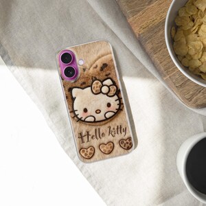 Może przedstawiać: Etui na telefon z motywem Hello Kitty na jasnobrązowym tle. Zawiera napis "Hello Kitty", r&oacute;żową osłonę obiektywu aparatu i trzy motywy serca na dole.