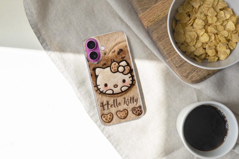 Może przedstawiać: Etui na telefon z motywem Hello Kitty na jasnobrązowym tle. Zawiera napis "Hello Kitty", r&oacute;żową osłonę obiektywu aparatu i trzy motywy serca na dole.