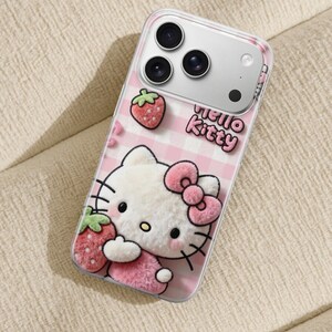 Może przedstawiać: Przezroczyste etui na telefon z motywem Hello Kitty w r&oacute;żowo-białą kratkę. Etui ma postać 3D Hello Kitty z r&oacute;żową kokardą i truskawkami. Na etui wydrukowano napis "hello kitty".