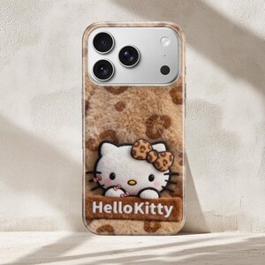 Może przedstawiać: Etui na telefon z motywem Hello Kitty. Etui ma brązową, puszystą fakturę z cętkami w panterkę. Postać Hello Kitty jest biała z kokardą i jest wyśrodkowana na etui, z napisem "Hello Kitty" poniżej.