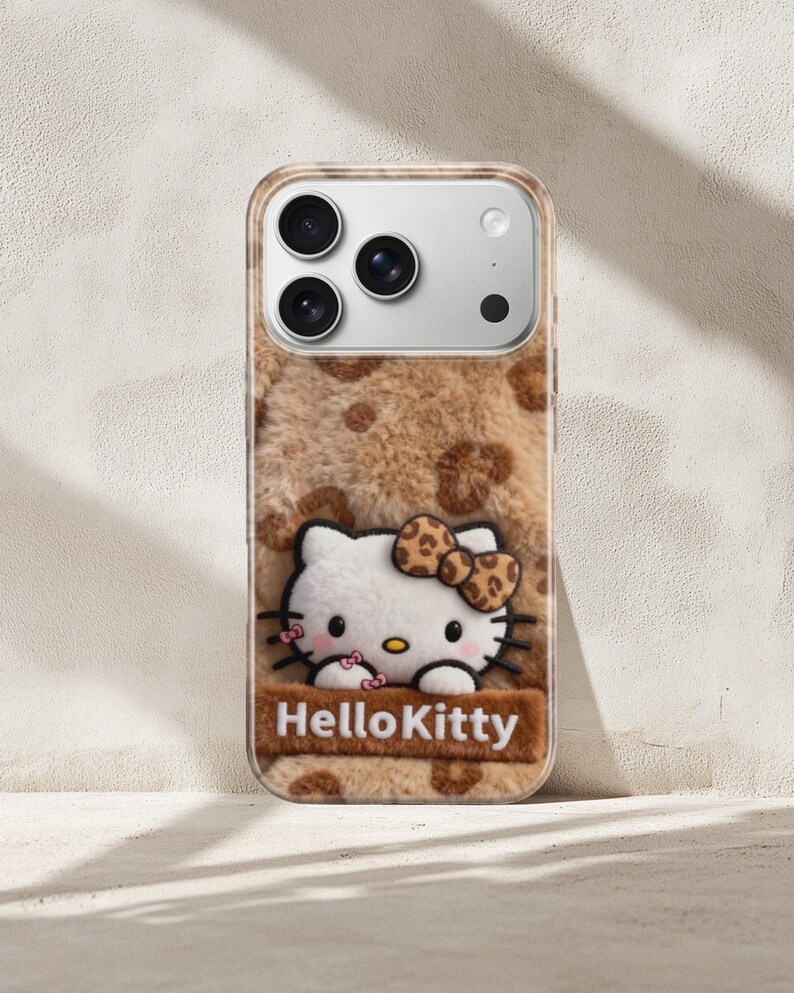 Może przedstawiać: Etui na telefon z motywem Hello Kitty. Etui ma brązową, puszystą fakturę z cętkami w panterkę. Postać Hello Kitty jest biała z kokardą i jest wyśrodkowana na etui, z napisem "Hello Kitty" poniżej.