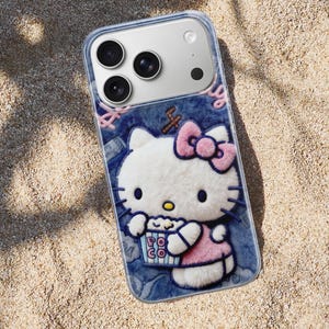Może przedstawiać: Przezroczyste etui na telefon z tr&oacute;jwymiarowym wzorem Hello Kitty trzymającej popcorn. Etui ma dżinsowe tło z r&oacute;żowymi akcentami i r&oacute;żową kokardą. Etui na telefon leży na piaszczystej powierzchni.