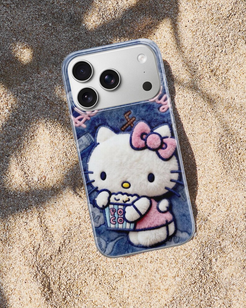 Może przedstawiać: Przezroczyste etui na telefon z tr&oacute;jwymiarowym wzorem Hello Kitty trzymającej popcorn. Etui ma dżinsowe tło z r&oacute;żowymi akcentami i r&oacute;żową kokardą. Etui na telefon leży na piaszczystej powierzchni.