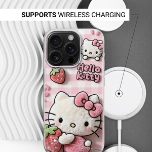 Może przedstawiać: Przezroczyste etui na telefon z motywem Hello Kitty na r&oacute;żowym tle w kratkę. Etui ma efekt 3D z Hello Kitty trzymającą truskawkę. Widoczny jest napis "Hello Kitty". Obsługuje ładowanie bezprzewodowe.