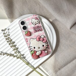 Może przedstawiać: R&oacute;żowe etui na telefon z Hello Kitty, truskawką i kokardą. Etui ma r&oacute;żowo-biały wz&oacute;r w kratkę i napis "Hello Kitty". Etui leży na białym talerzu.