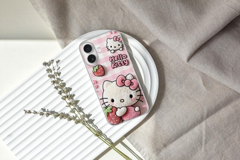 Może przedstawiać: R&oacute;żowe etui na telefon z Hello Kitty, truskawką i kokardą. Etui ma r&oacute;żowo-biały wz&oacute;r w kratkę i napis "Hello Kitty". Etui leży na białym talerzu.