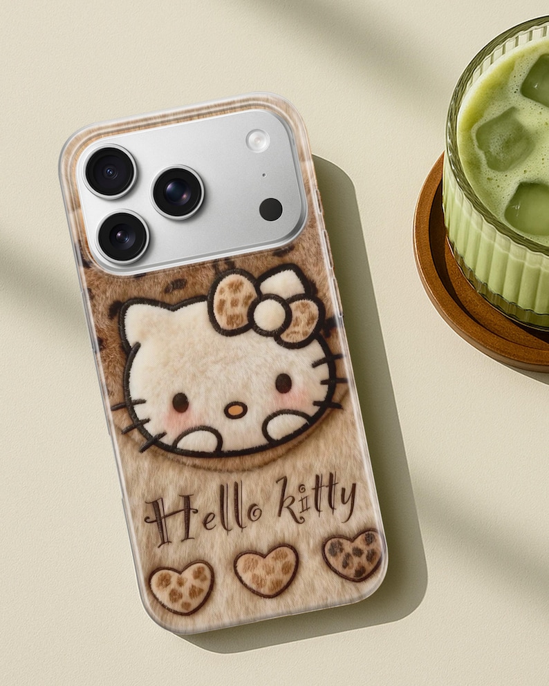 Może przedstawiać: Etui na telefon z motywem Hello Kitty. Etui ma brązową i beżową teksturę przypominającą futro z postacią Hello Kitty pośrodku. Słowa "Hello Kitty" są napisane pod postacią, wraz z trzema kształtami serca.