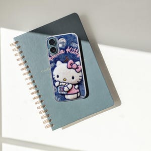 Może przedstawiać: Niebieskie etui na telefon z motywem Hello Kitty, r&oacute;żową kokardą i popcornem. Leży na niebieskim notatniku w spiralę ze złotą oprawą. Na etui widnieje napis "Hello Kitty".