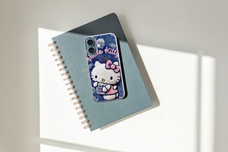 Może przedstawiać: Niebieskie etui na telefon z motywem Hello Kitty, r&oacute;żową kokardą i popcornem. Leży na niebieskim notatniku w spiralę ze złotą oprawą. Na etui widnieje napis "Hello Kitty".