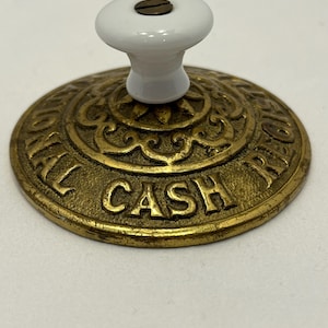 May include: A vintage brass cash register knob with a white ceramic handle. The knob is circular with ornate detailing and the word "CASH" is prominently displayed. The brass has a warm, aged patina.