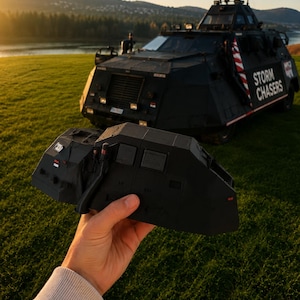 以下が含まれることがあります： 手前に持たれた黒く角張った3Dプリント製の車両ルーフセクションの模型と、背景の草地に駐車された「STORM CHASERS」と書かれた実物大の黒い装甲車両。夕暮れ時の空の下で撮影されています。