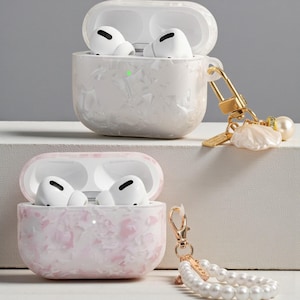 Può includere: Due set di auricolari wireless bianchi in custodie decorative. Una custodia è bianca con finitura perlata, l'altra è rosa con un design marmorizzato. Entrambe le custodie sono aperte, rivelando gli auricolari. Ogni custodia ha un portachiavi color oro.