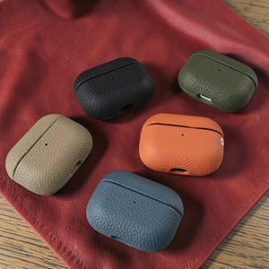 Puede incluir: Cinco fundas de cuero para AirPod en varios colores: negro, verde oliva, beige, naranja y azul. Las fundas tienen una superficie texturizada y están dispuestas sobre una superficie roja. Estas fundas están diseñadas para proteger y transportar auriculares inalámbricos.