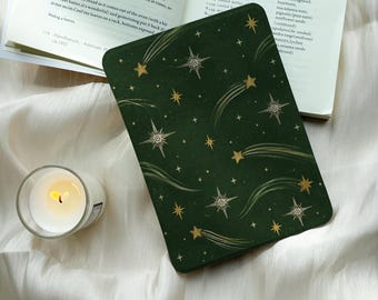 Può includere: Una custodia per e-reader verde scuro con un motivo di stelle e stelle cadenti dorate e bianche. Un libro di cucina aperto con testo e una piccola candela in vetro con fiamma accesa sono posizionati accanto alla custodia su un tessuto bianco.