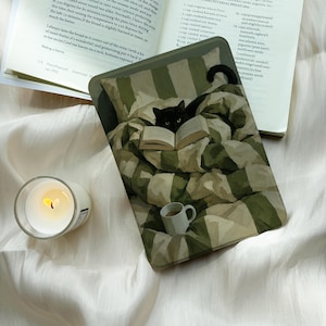 May include: A rectangular e-reader case featuring an illustration of a black cat nestled in a green and white striped blanket, reading an open book. A small white mug sits near the bottom edge of the case, which is placed next to an open cookbook.