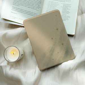 May include: A beige e-reader cover featuring a subtle watercolor-style sage green wash and gold foil starburst accents, positioned next to an open cookbook and a small glass candle holder with a lit white wax candle.