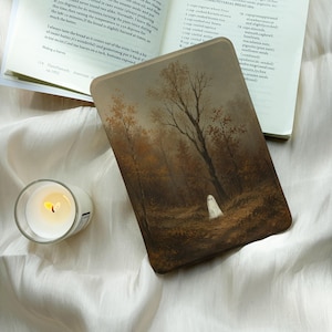 May include: A rectangular e-reader case featuring a dark, moody forest painting with brown and orange autumn trees and a small white ghost figure. Beside the case is a small glass candle holder with a lit white candle and an open cookbook.