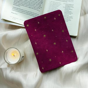 May include: A deep magenta e-reader cover featuring a scattered pattern of gold star icons, positioned next to an open cookbook with printed text and a small glass candle holder containing a lit white wax candle.
