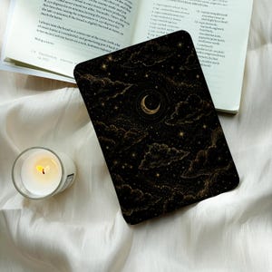 May include: A rectangular e-reader cover featuring a black background with gold celestial illustrations of a crescent moon, scattered stars, and clouds. The cover is placed next to an open cookbook and a small glass candle holder.