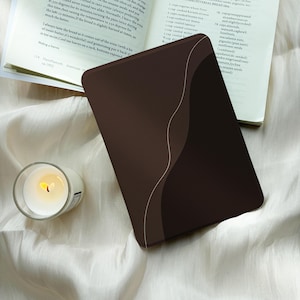 May include: A rectangular e-reader device featuring a dark brown and black abstract design with a thin, light-colored wavy line separating the two colors. The device is positioned on a white fabric surface next to an open book and a small candle.